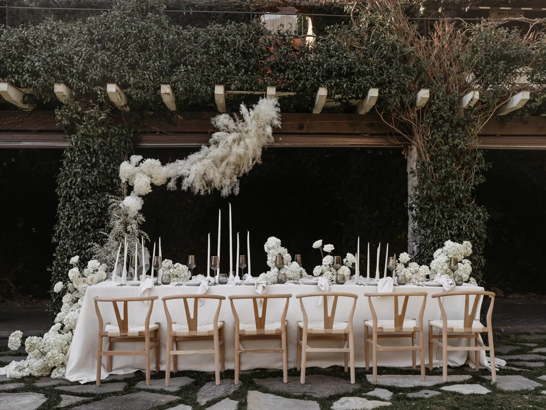 Hochzeitstafel unter einer begrünten Pergola mit Pampasgras, weißer Floristik und Holzstühlen
